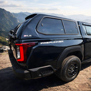 A black PreRunner S-JS pickup truck with a hard canopy cab is parked on a rocky mountain cliffside, with layered mountain ranges in the background under a hazy sky.